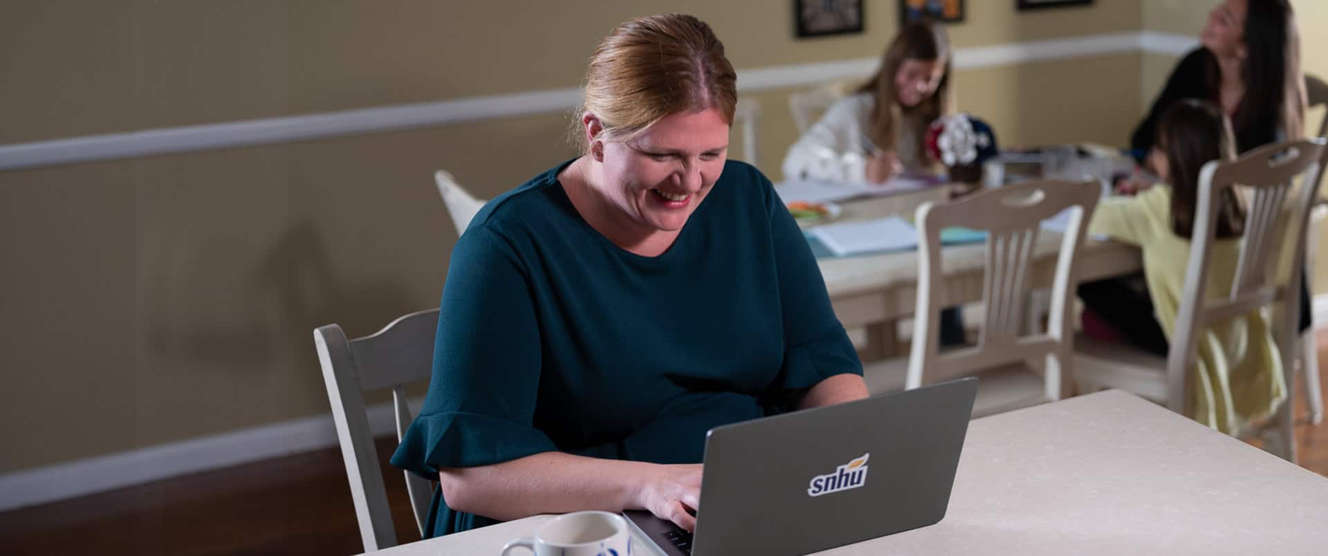 Shelly Villa, who earned a degree in criminal justice 2019, smiling and working on her laptop with two girls sitting at a table behind her.