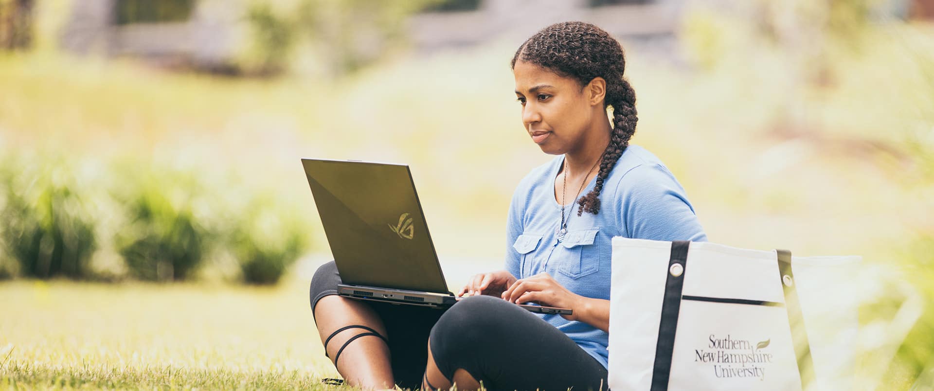"Bethanie Thomas, who earned their degree from SNHU in 2021, sitting cross-legged in the  grass working on her laptop with a Southern New Hampshire University canvas bag on the  ground beside her.