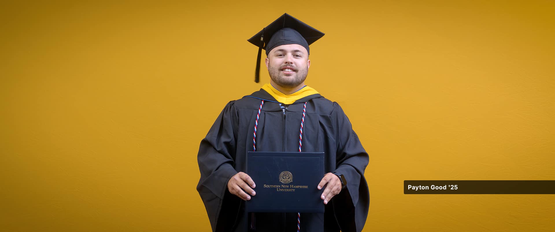 Payton Good, who earned his bachelor's in operations management from SNHU in 2025, wearing his cap and gown and holding his diploma in front of a yellow backdrop.