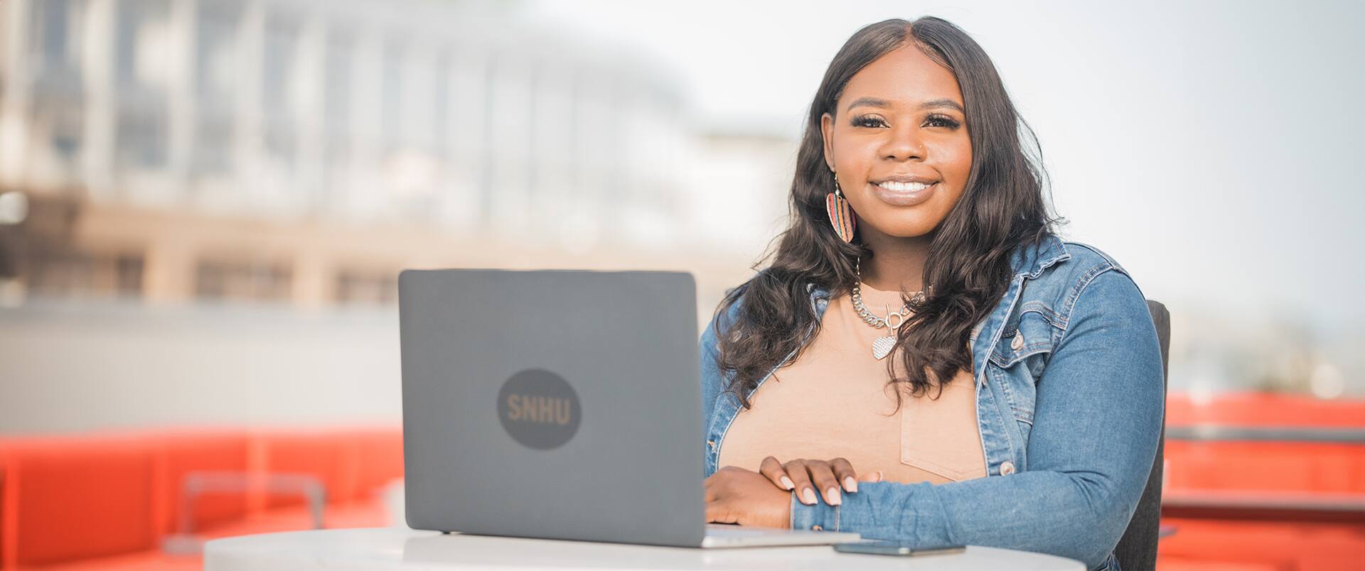 Tanzania Fair, who earned her degree from SNHU in 2020, wearing a denmin jacket and sitting at a small talbe with a laptop that has an SNHU sticker in front of her.