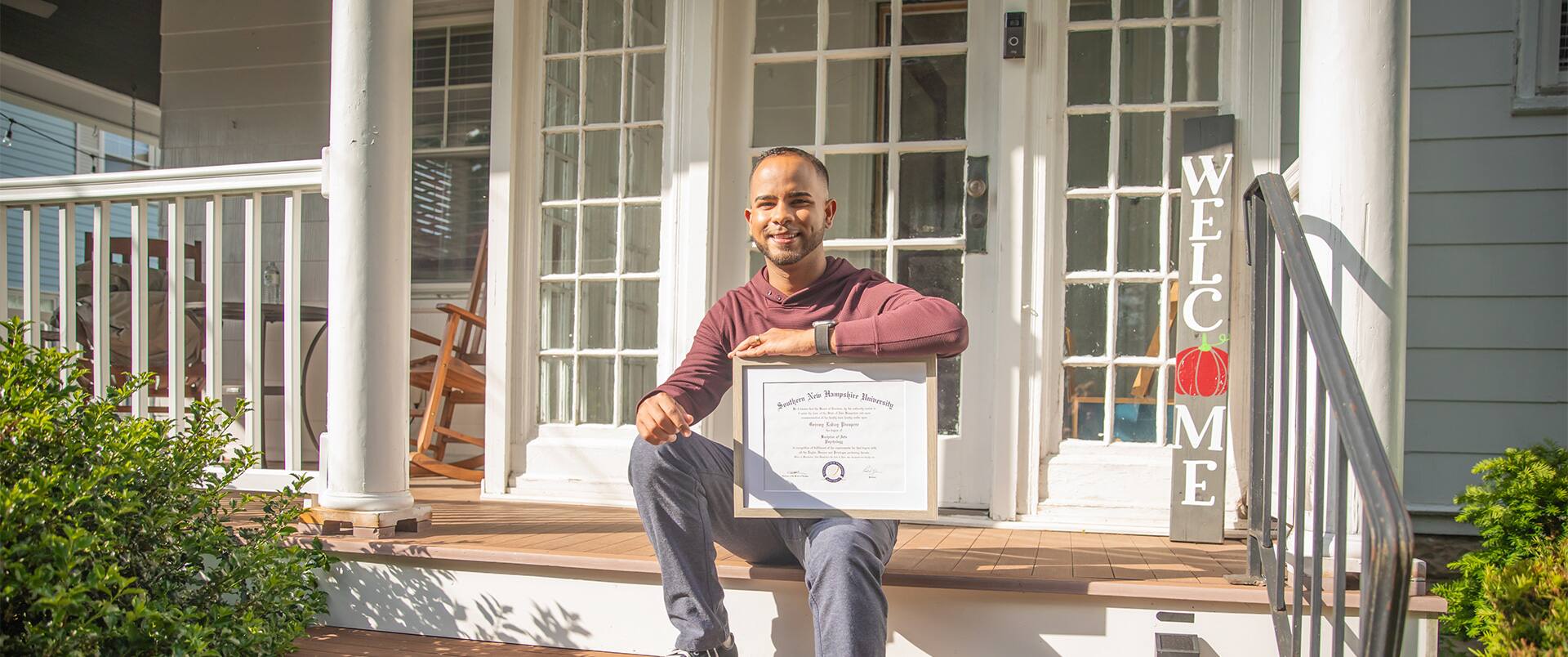 Geremy Laboy, who earned his degree from SNHU, sitting on a porch step holding his framed diploma on his knee with a vertical welcome sign in the background.
