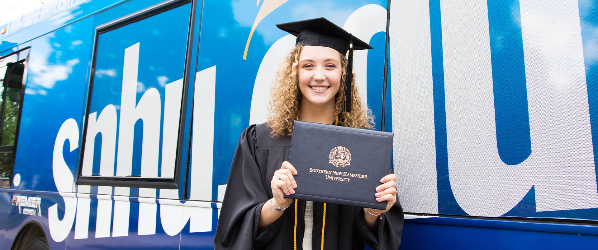 Joanne Coffey, wearing her cap and gown and holding her diploma while standing in front of the SNHU-branded bus.