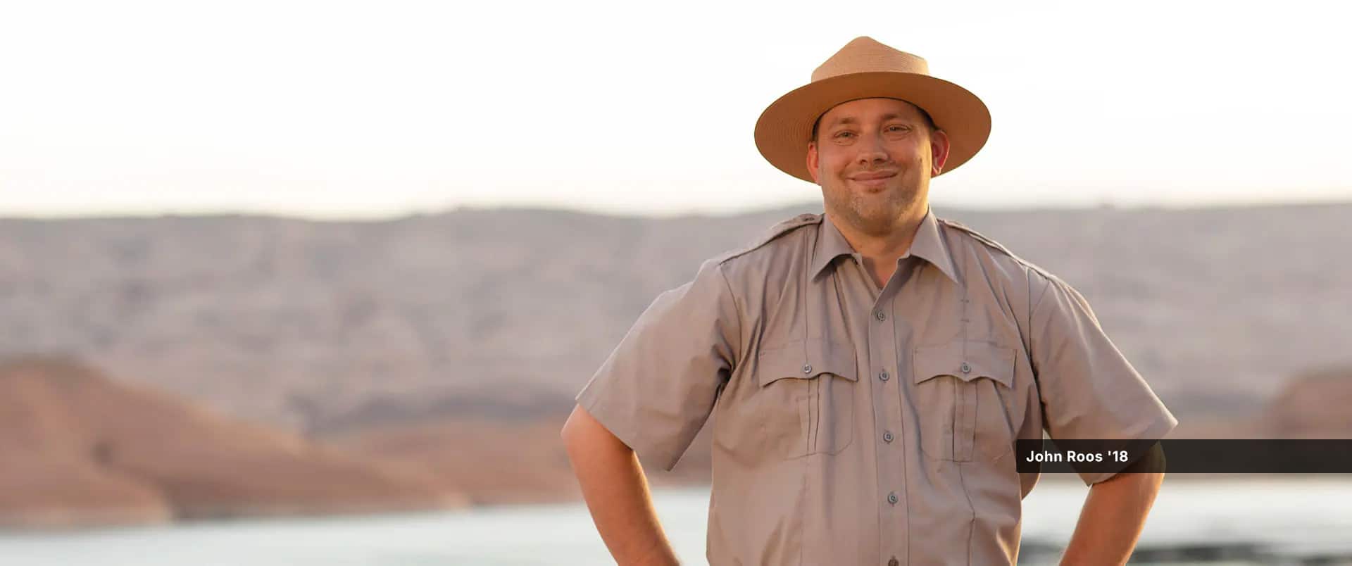 John Roos, who earned his online history degree from SNHU in 2018, wearing a tan park ranger uniform and hat, standing with his hands on his hips with a lake and low mountains visible in the background.