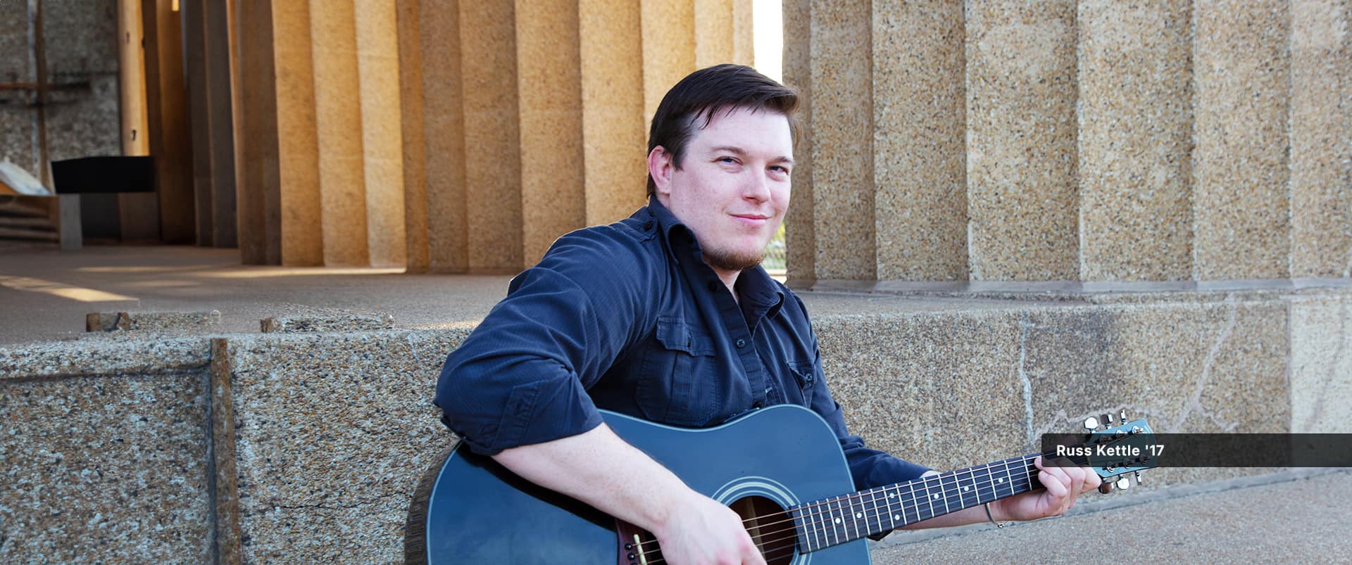 Russ Kettle, who earned his degree from SNHU in 2017, sitting on a set of stone steps playing a black acoustic guitar.