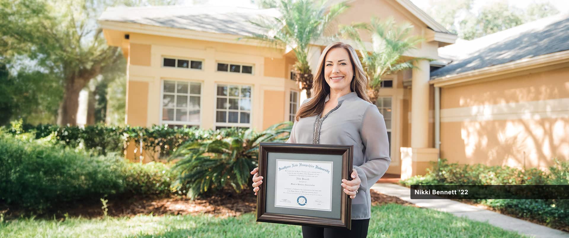Nikki Bennett, who earned her online MBA from SNHU in 2022, standing in her front yard holding her framed diploma.