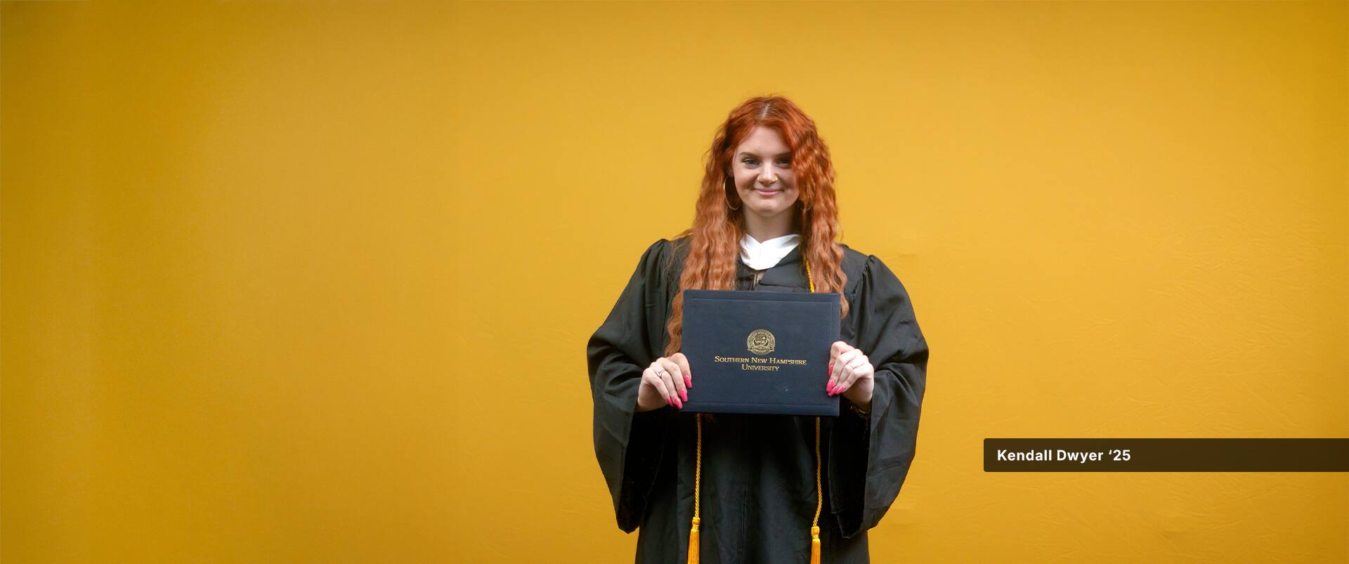 Kendall Dwyer, who earned her online education degree from SNHU in 2025, wearing her graduation robe and holding her diploma in front of a yellow backdrop.