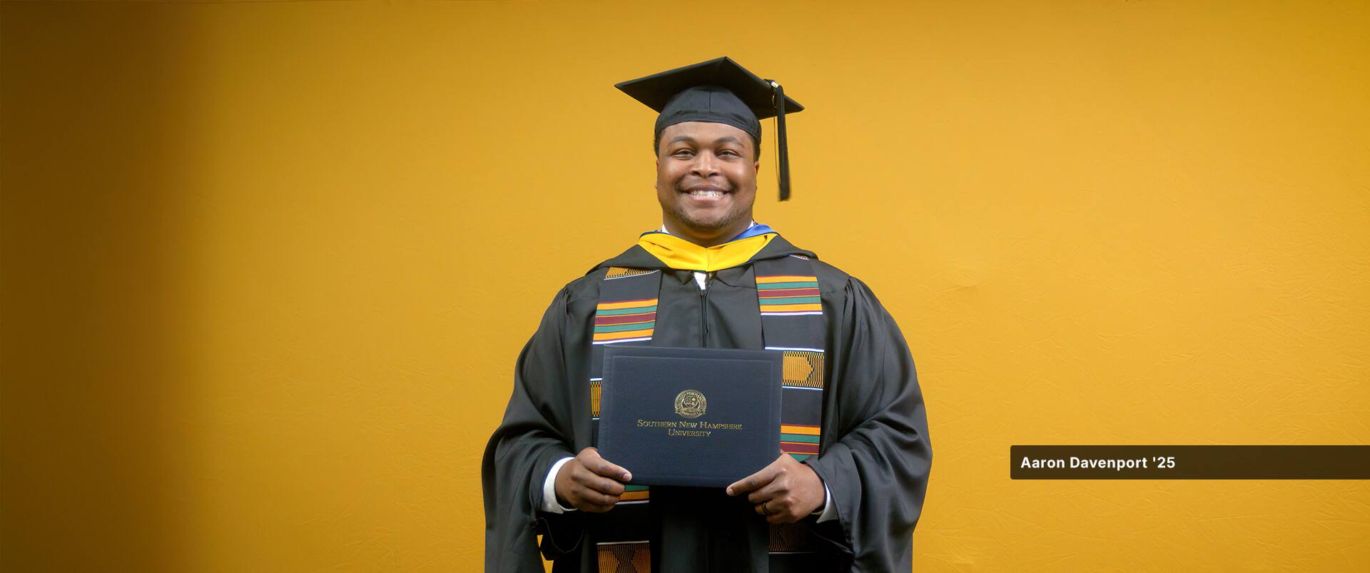Aaron Davenport, who earned his master's in finance from SNHU in 2025, wearing his cap and gown and holding his diploma in front of a yellow backdrop.