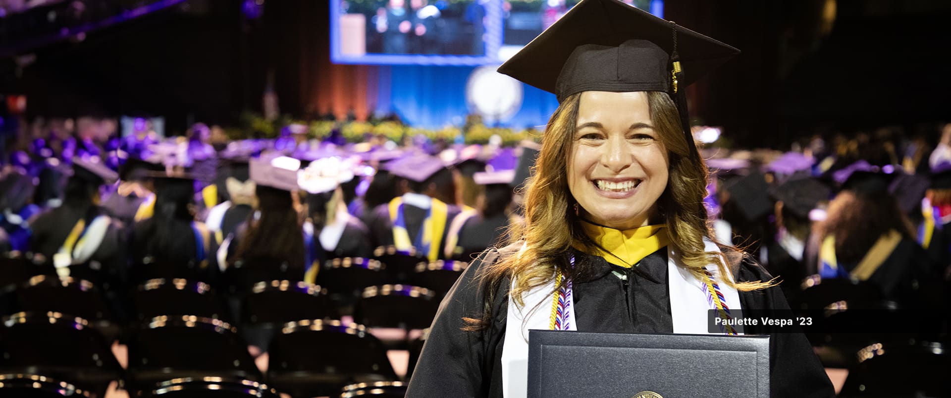 Paulette Vespa, who earned her master's in healthcare administration from SNHU in 2023, wearing her cap and gown and holding her diploma with a crowd of other graduates seated in the background.