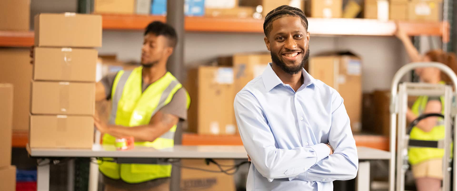 Matthew Seawright standing with his arms crossed in a shipping warehouse with someone behind him, stacking boxes.