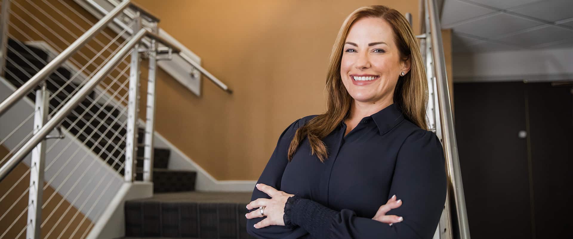 "Nikki Bennett, who earned her dgree at SNHU, wearing a dark blouse and standing with her arms folded  in a stairwell."
