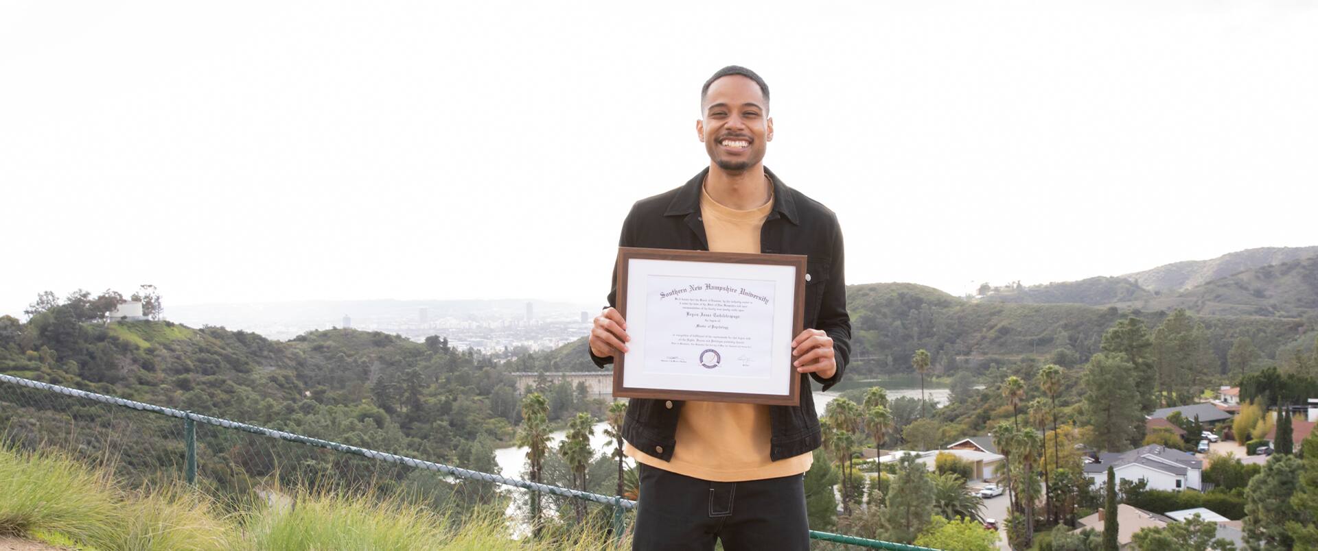 Keyon Tuitelelepaga, who earned his degree from SNHU in 2024, holding his framed diploma in front of him with houses, forested hills and palm trees in the background.