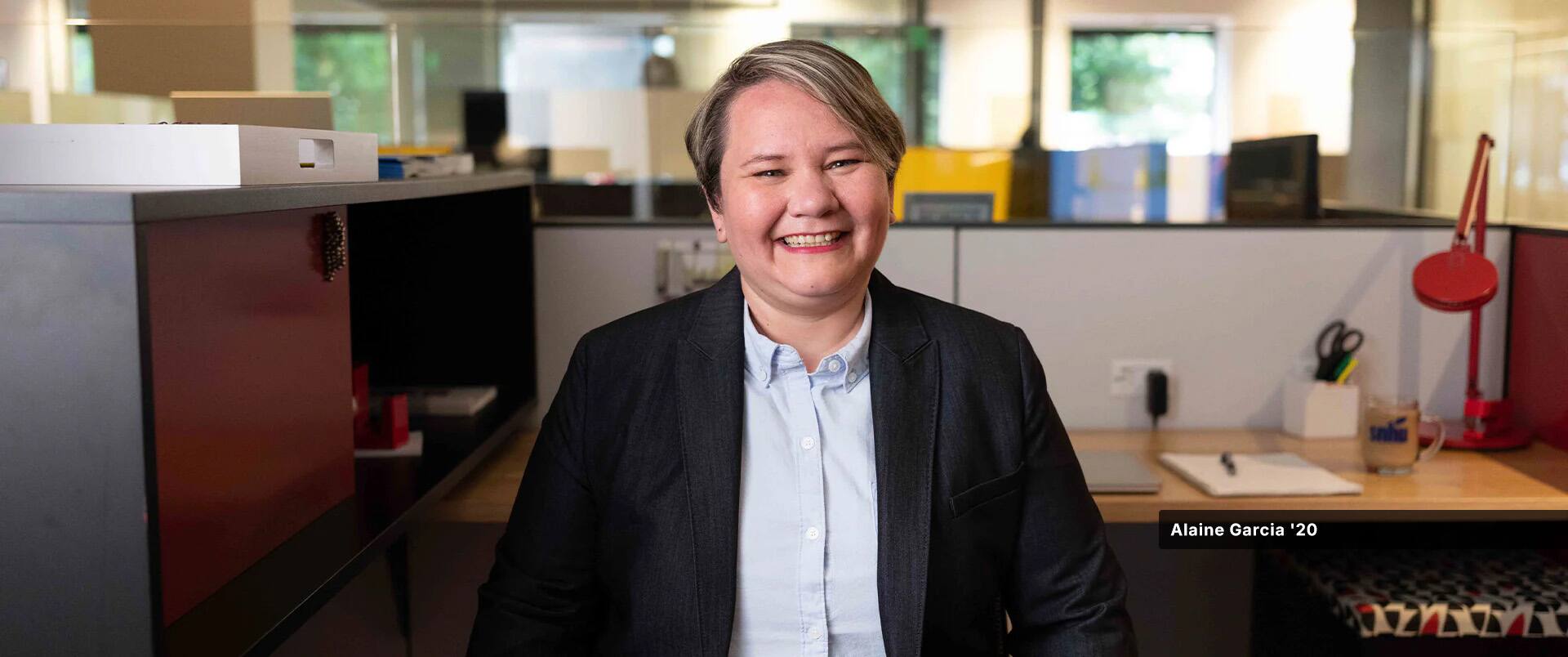 Alaine Garcia, who earned her degree from SNHU in 2020, wearing a dark blazer and blue button down  shirt standing in an office cubicle.