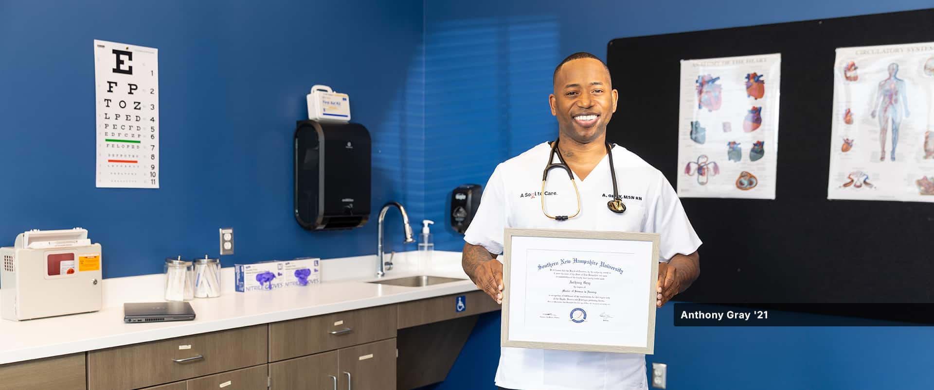 Anthony Gray, who earned his online master's in nursing from SNHU in 2021, standing in a doctor's office wearing white scrubs and a stethoscope around his neck and holding his framed diploma.