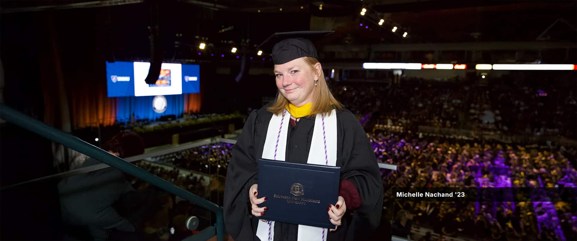 Michelle Nachand, who earned her bachelor's in nursing from SNHU, wearing her cap and gown and holding her diploma in a balcony above the commencement floor.