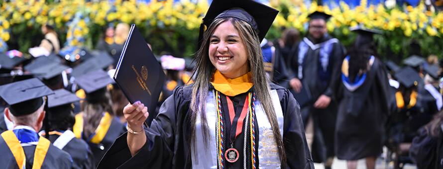 A smiling graduate at SNHU’s Spring 2026 commencement ceremony while holding their diploma, wearing a cap and gown with honors cords.