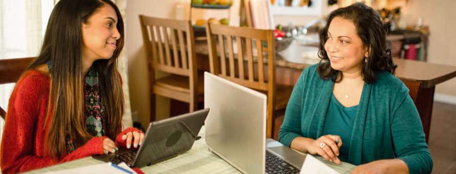A mother and daughter both on their laptops at a dining room table