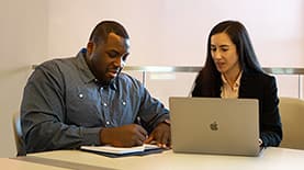 Two students working together, one writing in a notebook, one on a laptop