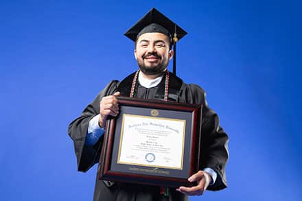 SNHU graduate, Jesus Suarez, standing in a cap and gown with his framed bachelor's degree