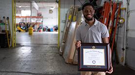 SNHU Graduate holding his framed diploma inside a warehouse workspace