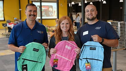 Two men and one woman holding colorful backpacks at an SNHU-sponsored Back to School Drive.