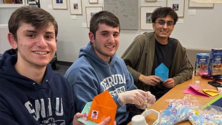 Three male students sitting at a table holding small colorful envelopes.