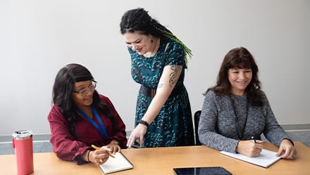 Two women sitting at a conference table with a third woman standing behind them and pointing at a notebook.