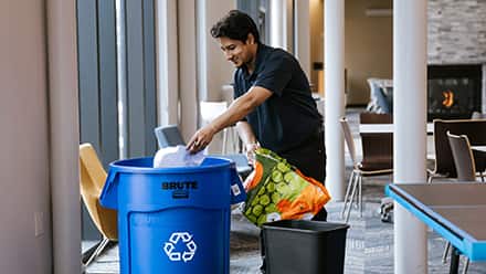 A man holding a reusable shopping bag putting a styrofoam container into a blue recycling barrel.