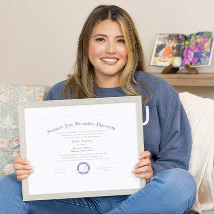 Juliana Zambrano, a 2025 SNHU graduate with a bachelor's degree in business administration sitting on a couch holding her diploma.
